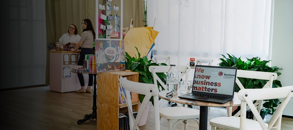 Interior view of a cozy cafe workspace featuring a laptop displaying Business Chamber Queensland, wooden chairs, and staff chatting in the background near a display setup.