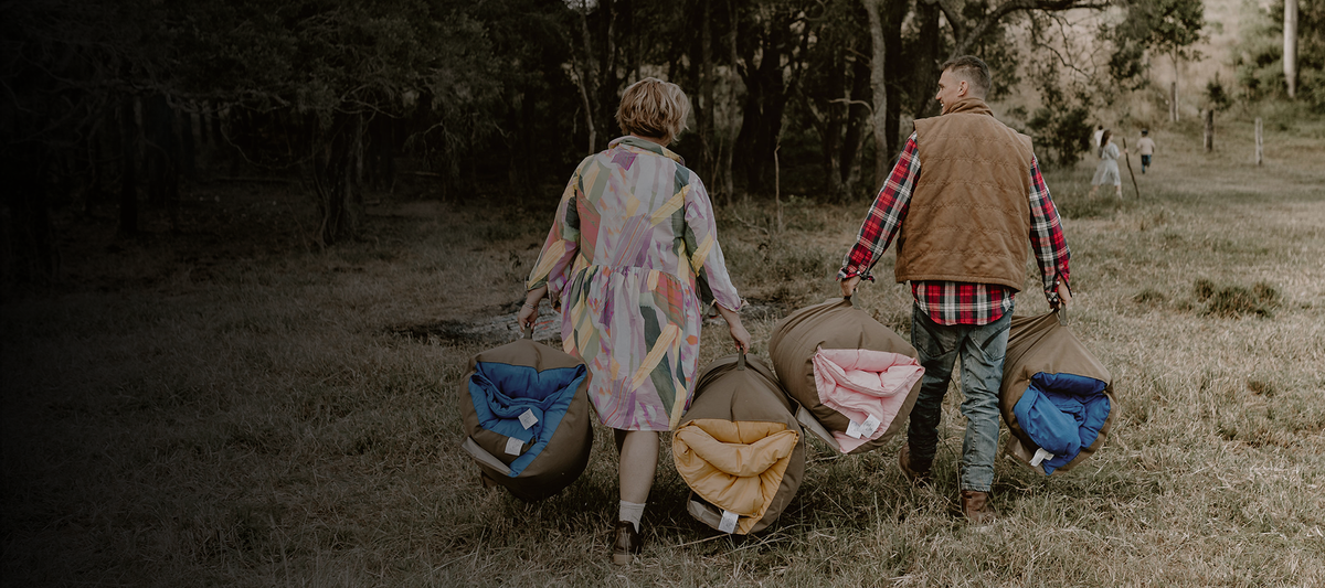 Cozie Dozie Co. founders walking through a grassy field carrying rolled Joey and Buck adventure loungers in BowerBird Blue, Ochre, and Quartz canvas carry bags.