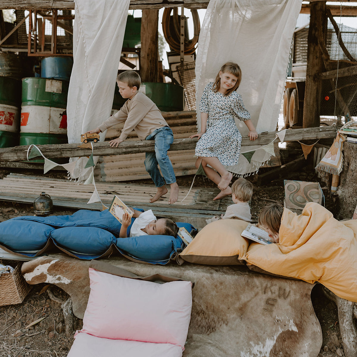 Children playing and reading on Cozie Dozie Co. Joey lounger covers in BowerBird Blue, Ochre, and Quartz inside a rustic country farm shed in Mount Larcom.