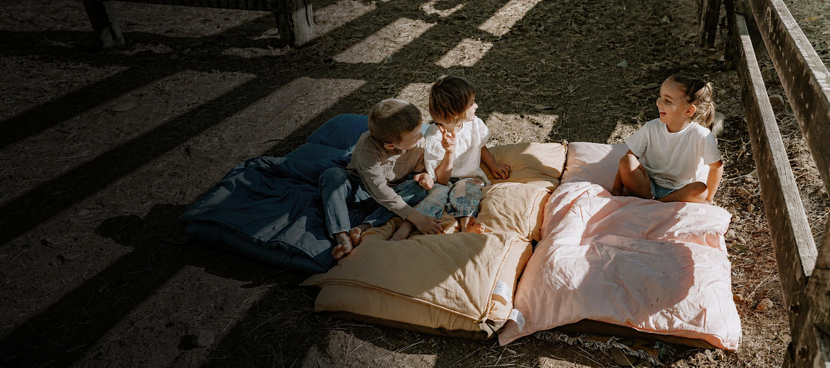 Three young children sitting together outdoors on Cozie Dozie Co. Joey pocket-style swag loungers in BowerBird Blue, Ochre, and Quartz.