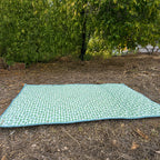 Quilted green gingham Co-mpanion Swag Mat with light blue trim laid flat on grass in an outdoor garden setting.