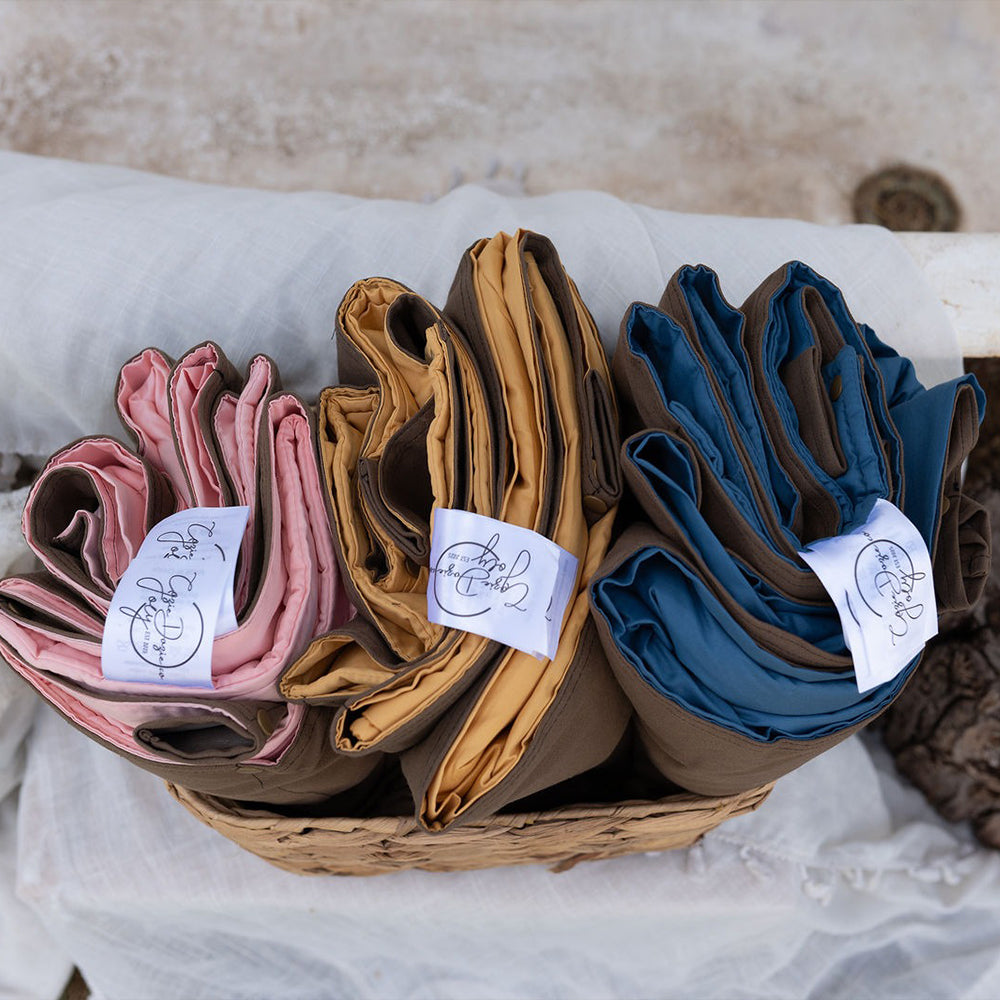 Top-down view of three neatly folded Cozie Dozie Co. Joey adventure loungers in Quartz pink, Ochre yellow, and BowerBird Blue placed inside a woven storage basket.
