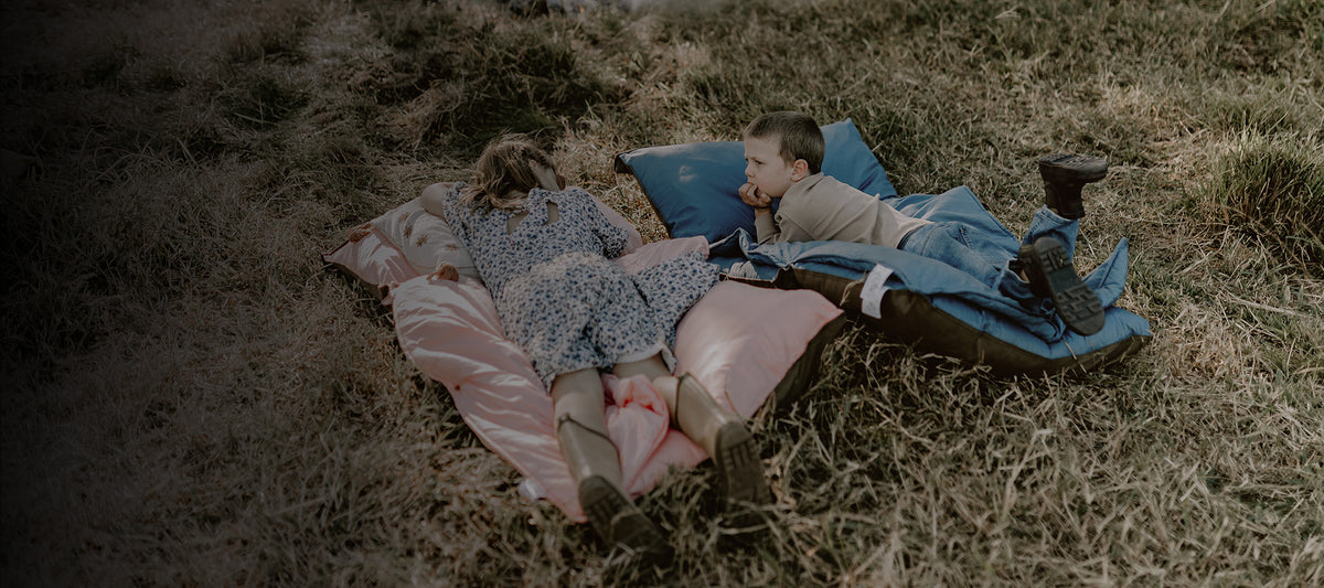 Two young children resting comfortably on Quartz pink and BowerBird Blue Joey adventure loungers in a grassy country field.