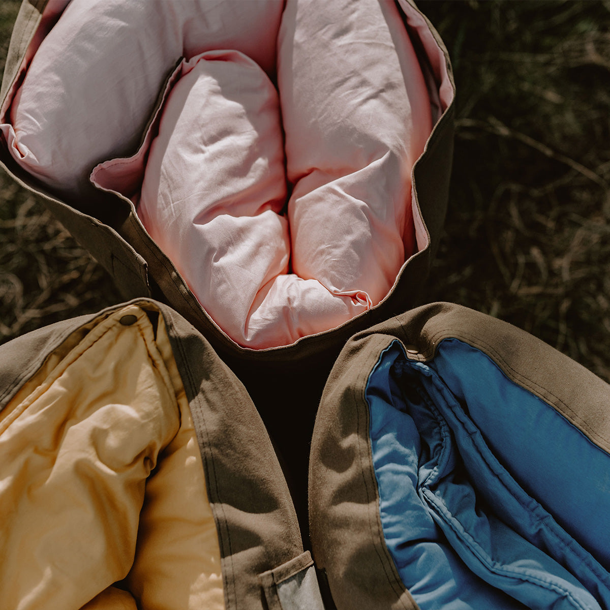Top-down view of three rolled Joey adventure loungers in Quartz pink, Ochre yellow, and BowerBird Blue resting together on dry country grass.