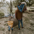 Father and young son walking through the bush near a campfire carrying rolled Cozie Dozie Co. Joey adventure loungers in BowerBird Blue and Ochre.