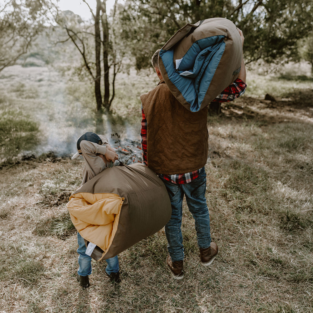Father and young son walking through the bush near a campfire carrying rolled Cozie Dozie Co. Joey adventure loungers in BowerBird Blue and Ochre.