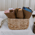 Front view of three folded Cozie Dozie Co. Joey adventure lounger covers in Quartz, Ochre, and BowerBird Blue neatly organized inside a natural woven basket.