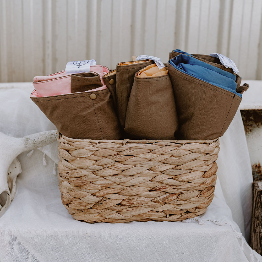 Front view of three folded Cozie Dozie Co. Joey adventure lounger covers in Quartz, Ochre, and BowerBird Blue neatly organized inside a natural woven basket.