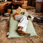 Person lying back reading a book on the Limited Edition Warrigal Greens gingham Joey adventure lounger inside a rustic hay barn.