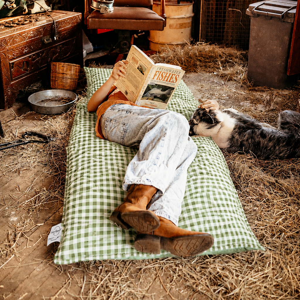 Person lying back reading a book on the Limited Edition Warrigal Greens gingham Joey adventure lounger inside a rustic hay barn.