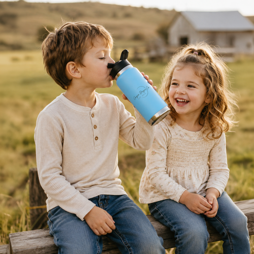 Young boy drinking water from a BowerBird Blue Cozie Dozie Co. stainless steel bottle while sitting on a wooden fence next to his laughing sister on a country farm.