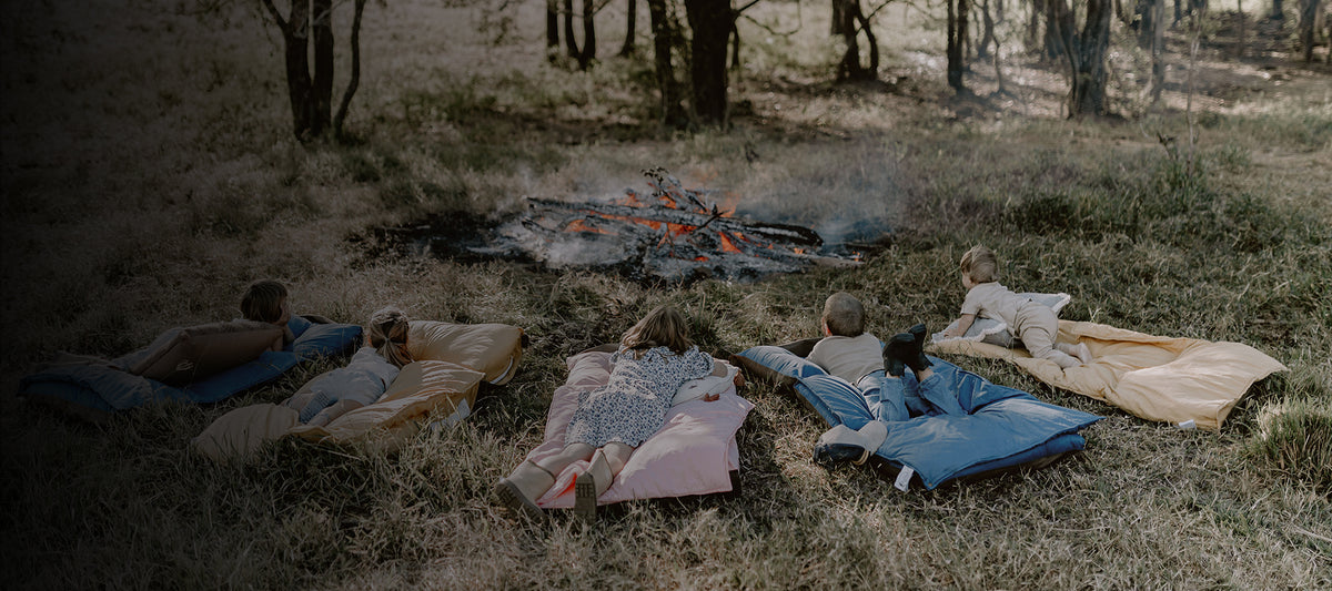 Group of five children lying stomach-down on Cozie Dozie Co. Joey adventure loungers in a row around an outdoor campfire in the Australian bush.