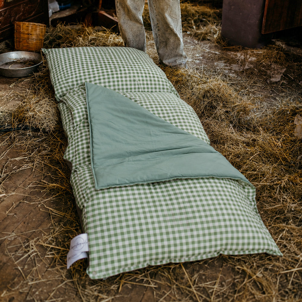 Filled Limited Edition Warrigal Greens Joey adventure lounger lying flat on a hay-covered barn floor, showing the green gingham print with its sage green French linen blanket peeled back.