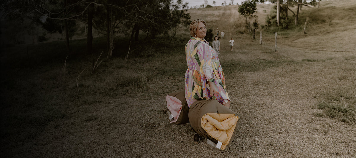 Mother in a colorful patterned dress carrying rolled Cozie Dozie Co. Joey adventure loungers along a grassy farm track with children following behind.
