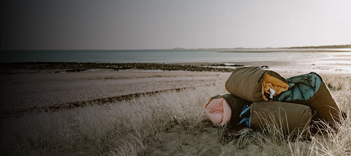 Rolled Cozie Dozie Co. Joey adventure loungers stacked in coastal beach grass overlooking the Central Queensland ocean at sunset.