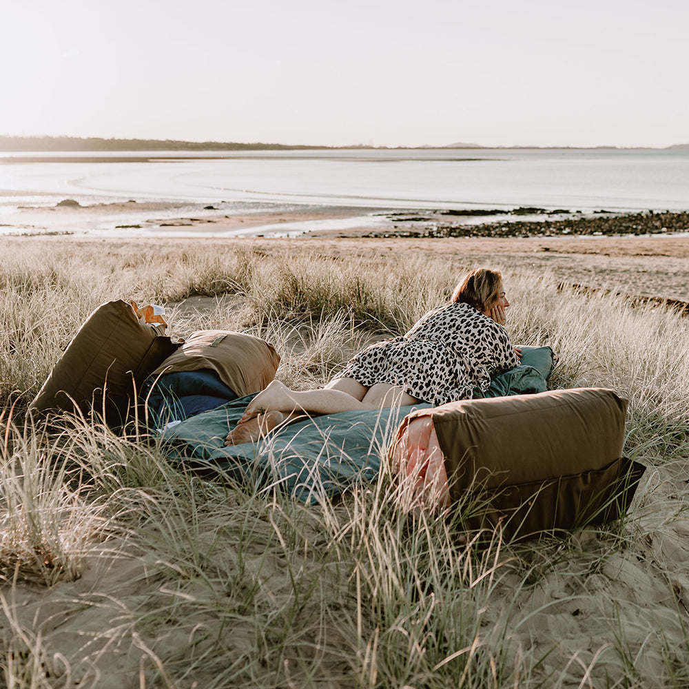 Woman relaxing on the extra-long Buck adventure lounger in Sea Moss green among coastal grass dunes overlooking the ocean.