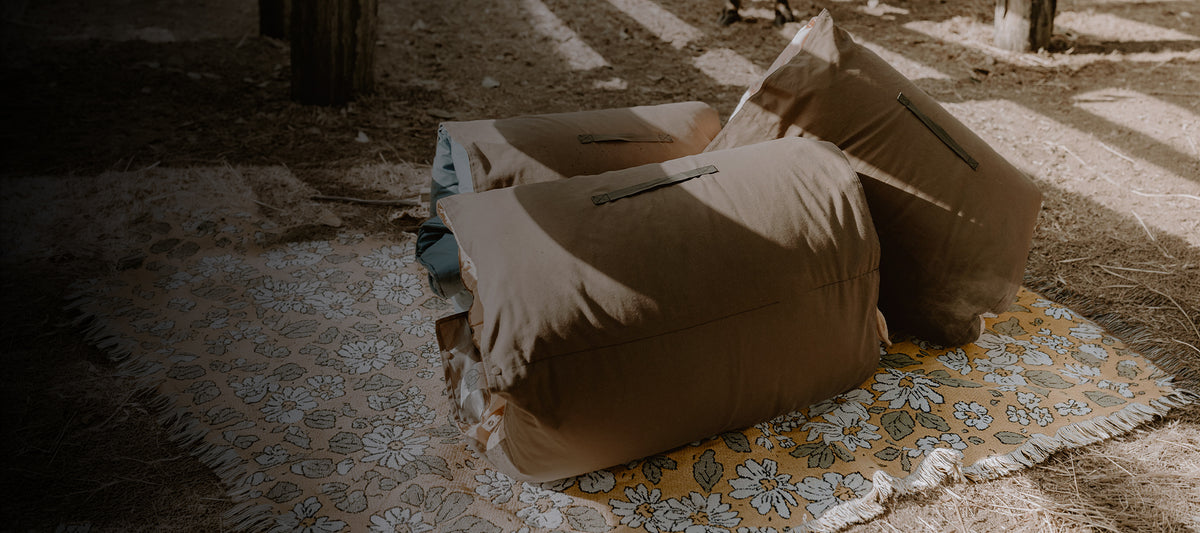Three rolled Cozie Dozie Co. adventure loungers showing canvas backing and carrying handles resting on a floral vintage picnic rug in a rustic shed setting.