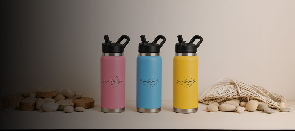 Three Cozie Dozie Co. 27oz insulated stainless steel drink bottles in Quartz pink, BowerBird Blue, and Ochre yellow arranged on a tabletop with river stones and a net mesh bag.