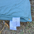 Close-up shot of the Sea Moss French linen corner, brass press stud, and Cozie Dozie Co. Buck product tag resting on outdoor grass.