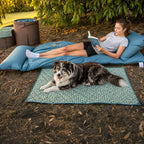 Teenager relaxing and reading a book on The Buck 3-metre double-length adventure lounger in Sea Moss while a dog lies beside her on a green gingham mat.