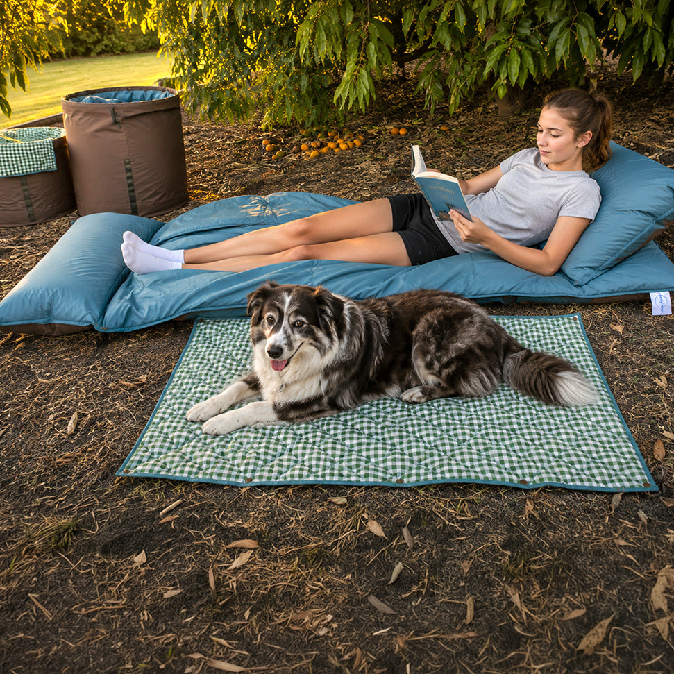 Teenager relaxing and reading a book on The Buck 3-metre double-length adventure lounger in Sea Moss while a dog lies beside her on a green gingham mat.