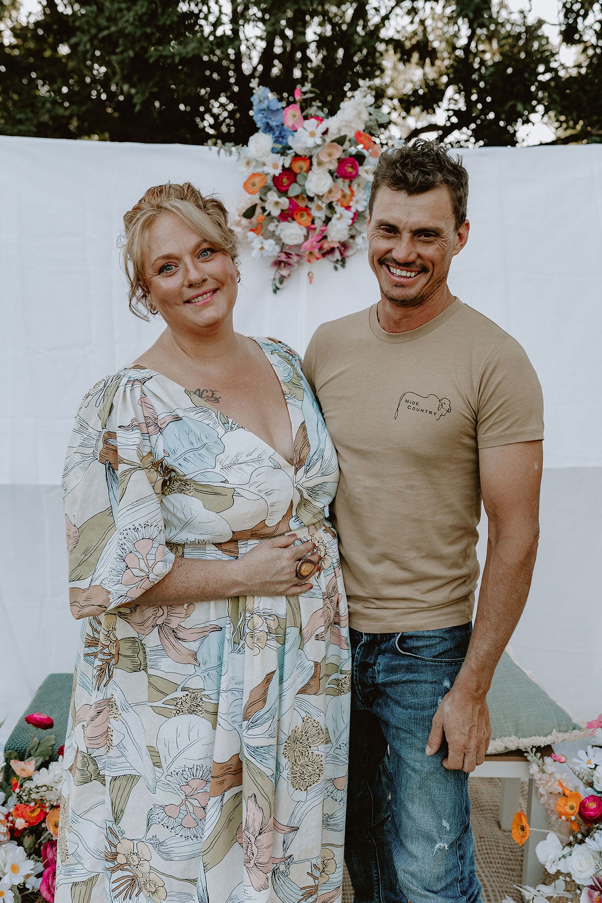 Cozie Dozie Co. founder Emma James-Skey standing next to her husband Sunny, smiling warmly in a rustic outdoor setting with native flowers behind them.